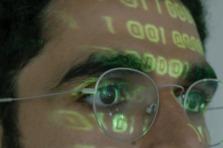 A close up picture of a person wearing spectacles with shadows of coding on his face.